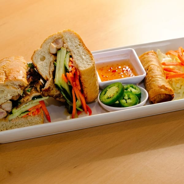 A Vietnamese banh mi sandwich, egg roll, and sides of sauce, jalapeños, and salad on a white plate.