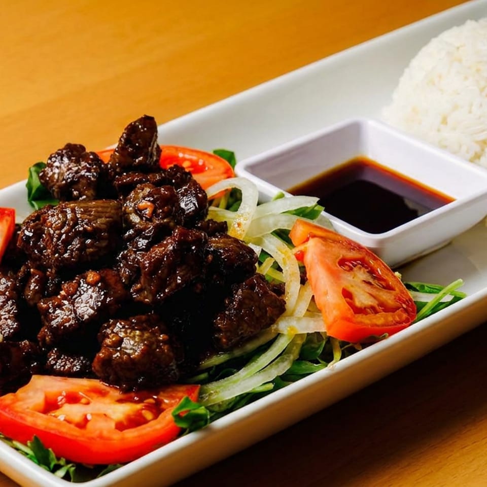 Shaking beef with rice, tomatoes, onions, and a side of sauce on a white rectangular plate.